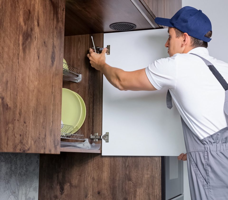 Male repair worker tightens bolts of cabinet with utensils using screwdriver in kitchen premise of apartment. Home repair service concept